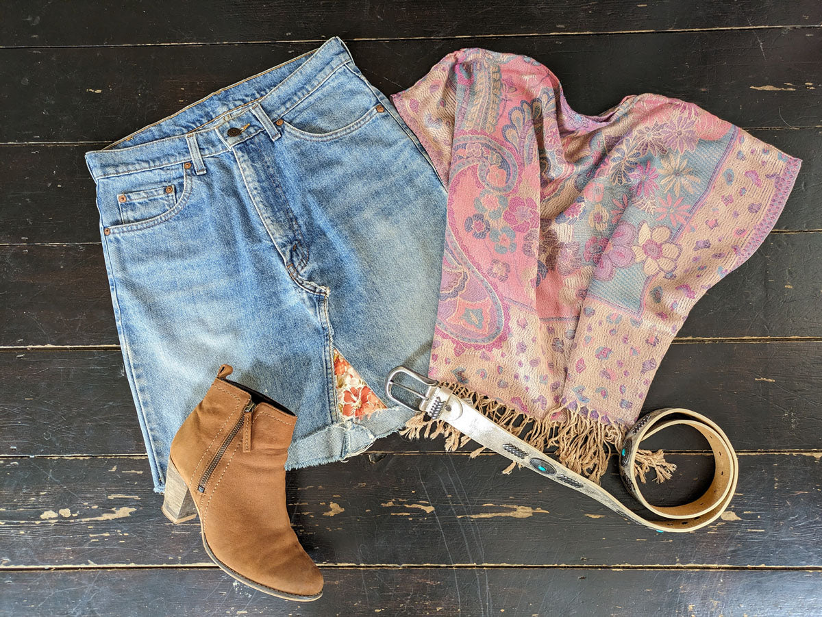 Folded jeans, brown boot, floral scarf, and belt on a dark wooden surface