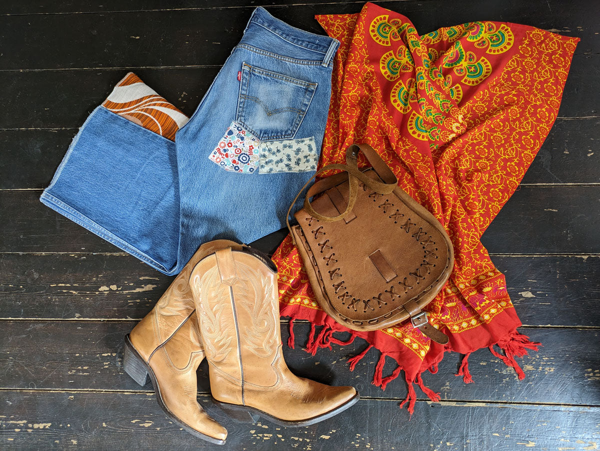 Blue jeans with floral pocket, brown leather boots, and a patterned red scarf on a dark wooden surface.