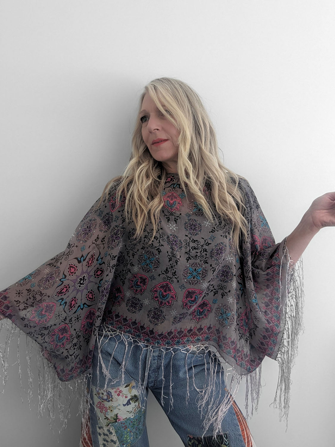 Person wearing a patterned poncho with fringe and jeans against a white background