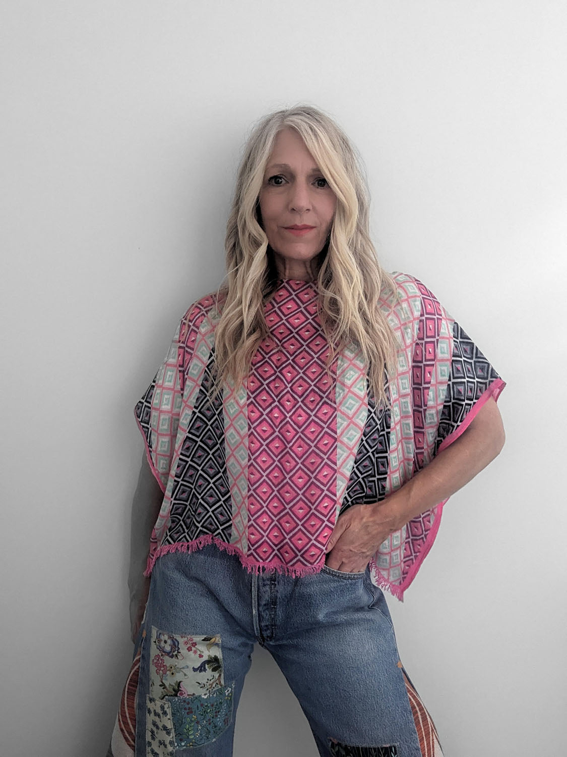 Woman wearing a patterned pink and black shawl over a floral top and jeans against a plain background
